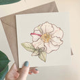 Load image into Gallery viewer, Querin Creations square greeting card with a camellia flower design held by a hand, with a brown envelope in the background.