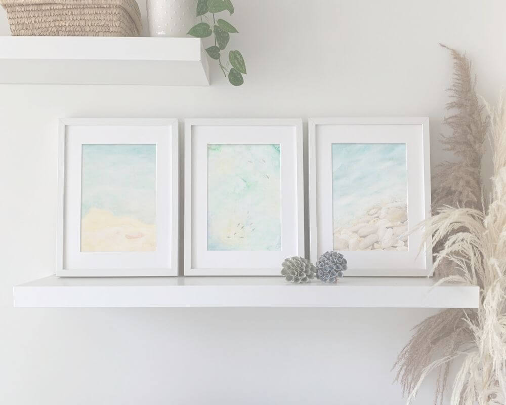 Three framed coastal artworks on a white shelf with decorative ceramics and dry leaves