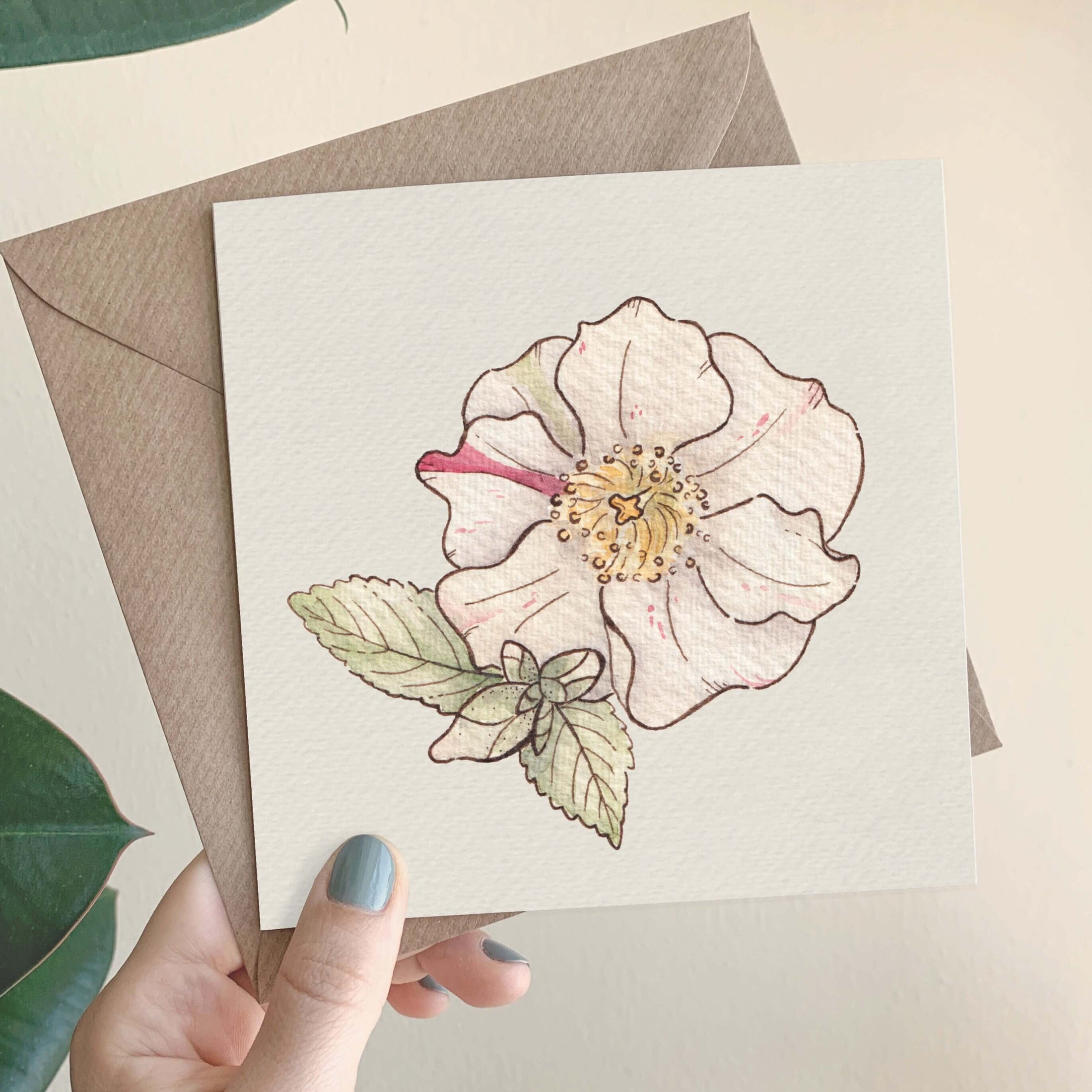 Querin Creations square greeting card with a camellia flower design held by a hand, with a brown envelope in the background.