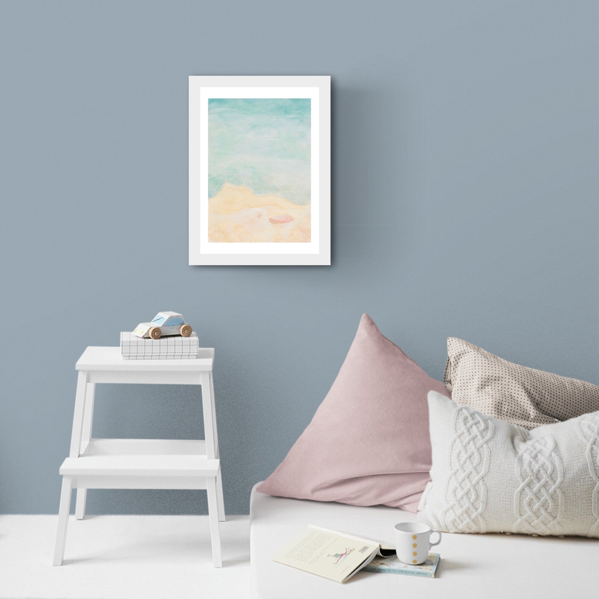 Querin Creations Framed seaside watercolour artwork on a light blue  wall with pillows and a small table below.