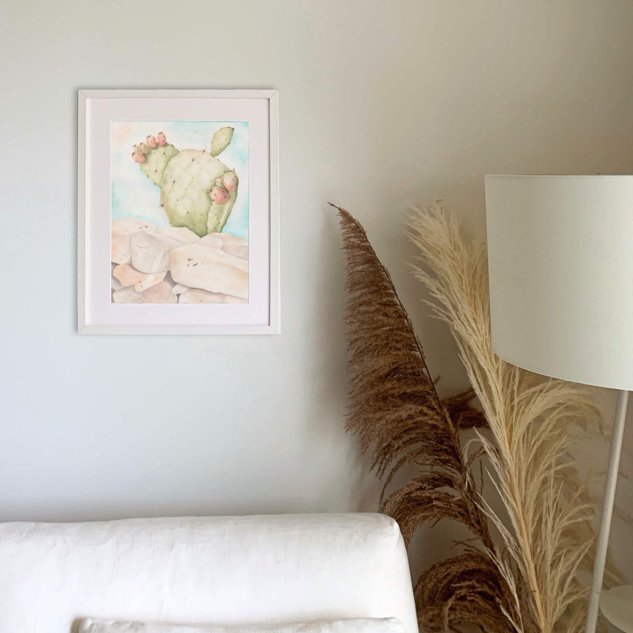 Querin Creations Framed watercolour artwork of a cactus on a wall with a lamp and decorative plant in the foreground above white sofa. Minimal, relaxing Home decor 