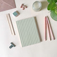 Load image into Gallery viewer, Desk setup with stationery items including a green stripes notebook, pens, and a candle on a light surface.
