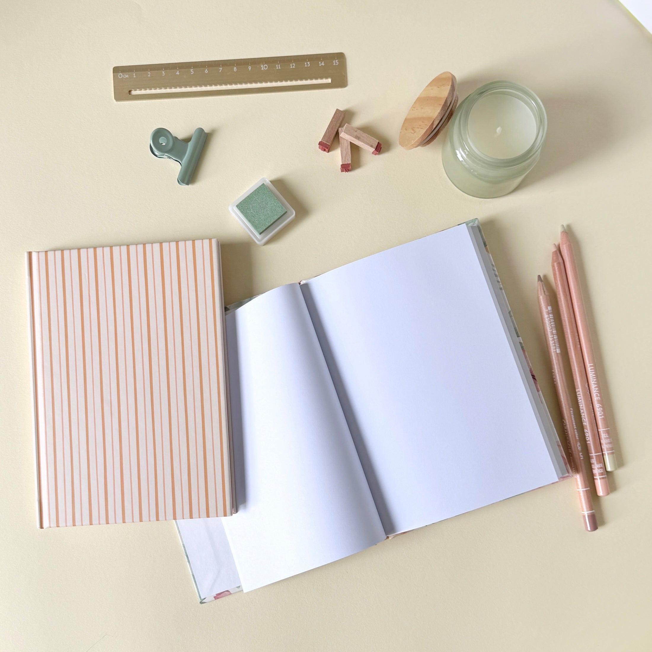 Open notebook with a yellow stripes notebook overlapped on a light desk with a candle and gold and green stationery items