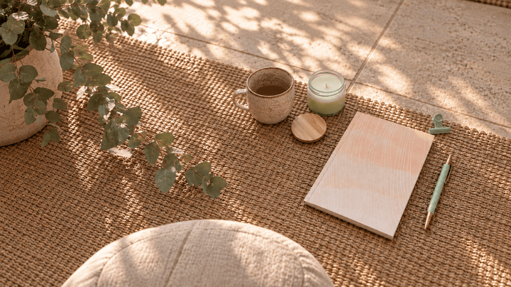 A styled flat-lay scene on a woven jute rug featuring a light pink shades notebook, a mint green pen, a small candle, a ceramic mug, and a potted plant, with dappled sunlight filtering through leaves.